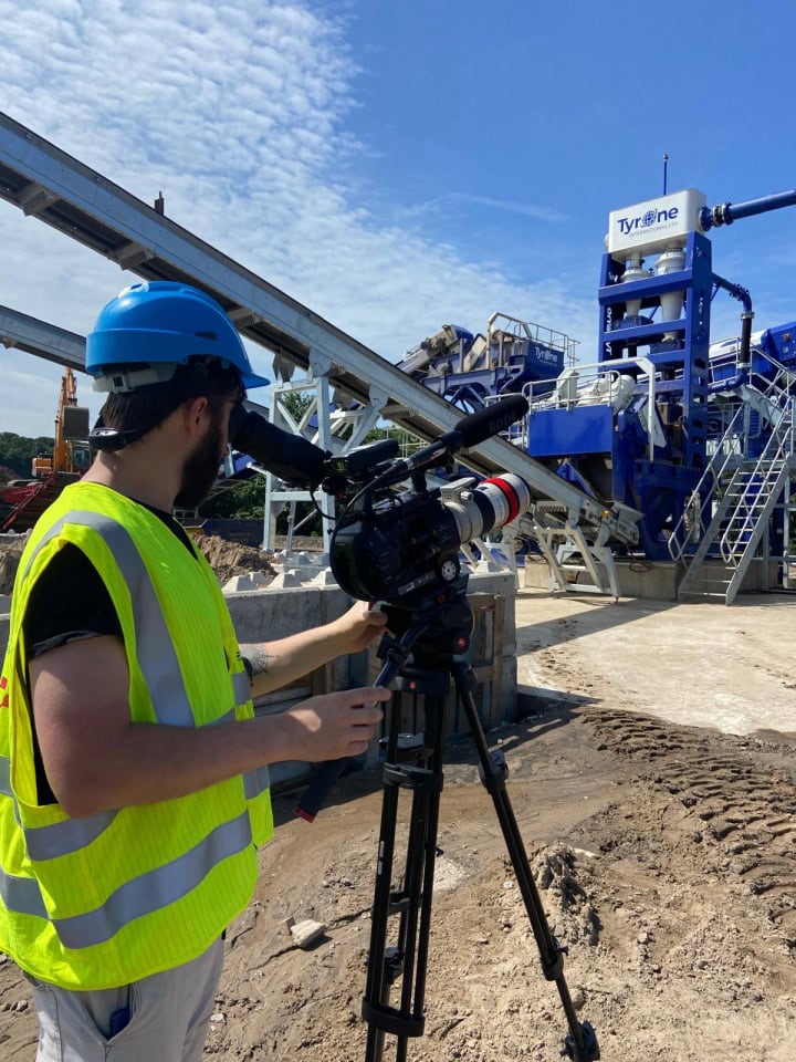 Cameraman filming at construction site with heavy machinery and conveyor belts.