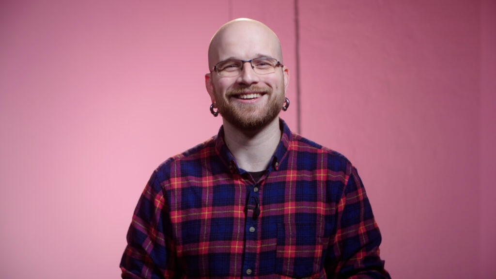 Friendly man with glasses, earrings, and a checkered shirt in a colourful studio setting. Fresh video content.